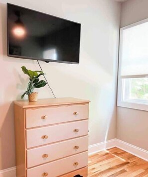 Une pièce dotée d’un téléviseur à écran plat fixé au mur, d’une commode en bois avec des tiroirs, d’une plante en pot et d’une fenêtre avec des stores.