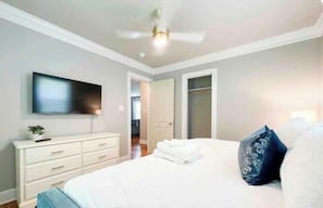 A bedroom with a bed, a TV mounted on the wall, a dresser, and a ceiling fan.