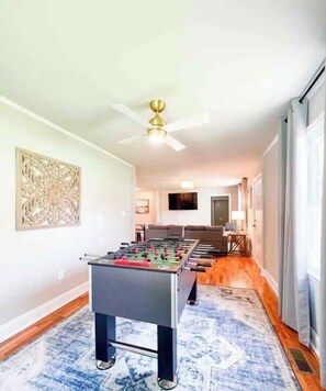 Un vaste espace de vie comprenant une table de baby-foot, un ventilateur de plafond et une grande œuvre d’art accrochée au mur.