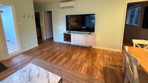Un'ampia zona giorno con TV a schermo piatto montata su un mobile bianco, pavimento in legno e un'ampia finestra che offre luce naturale.