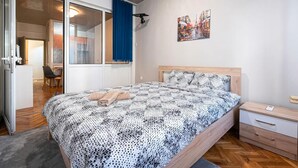 Une chambre à coucher avec un lit, une table de chevet, un bureau et une chaise. Il y a une fenêtre avec des rideaux et un tableau au mur.