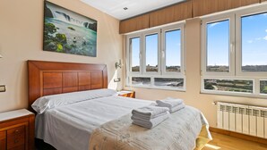 A bedroom with a large bed, a nightstand, a window with a view, and a painting on the wall. - Vacation Home 'Santa Divina Relax Para 10 Personas' with Balcony and Wi-Fi (Asturias)