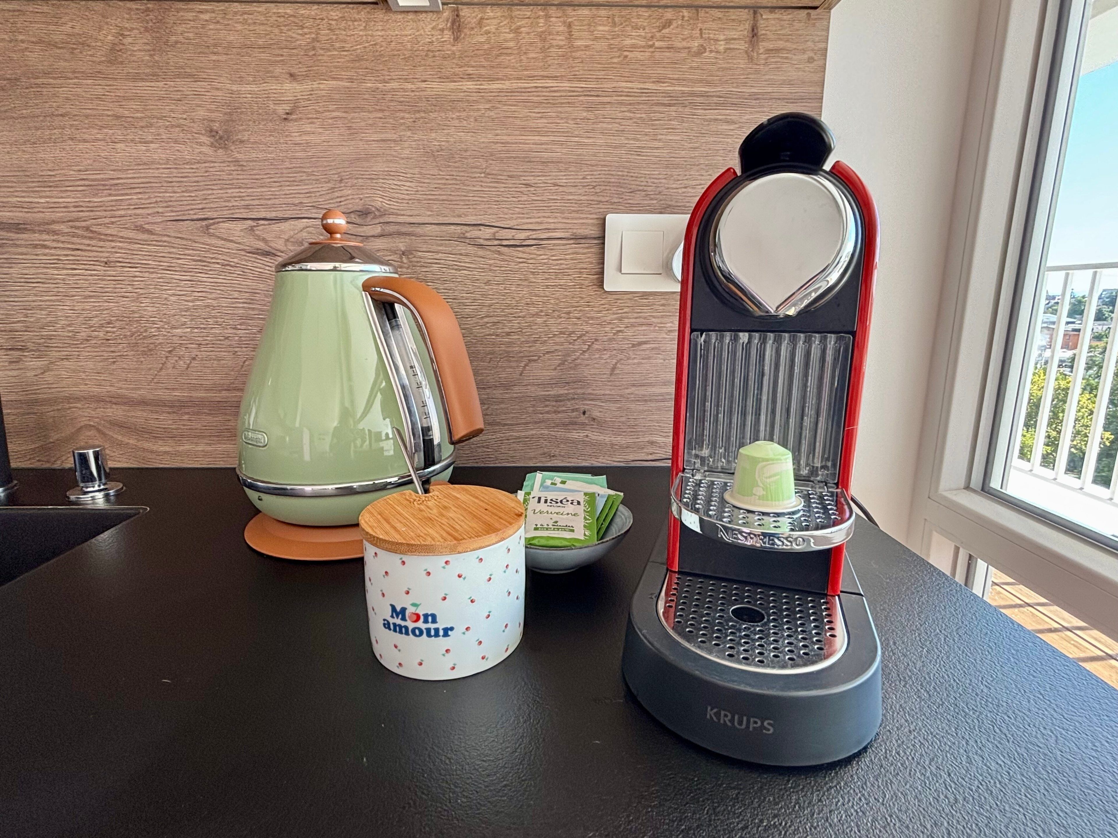Un plan de travail de cuisine avec une bouilloire verte, une tasse blanche portant l’inscription « Mon amour », un moulin à café Krups et une boîte de sachets de thé.