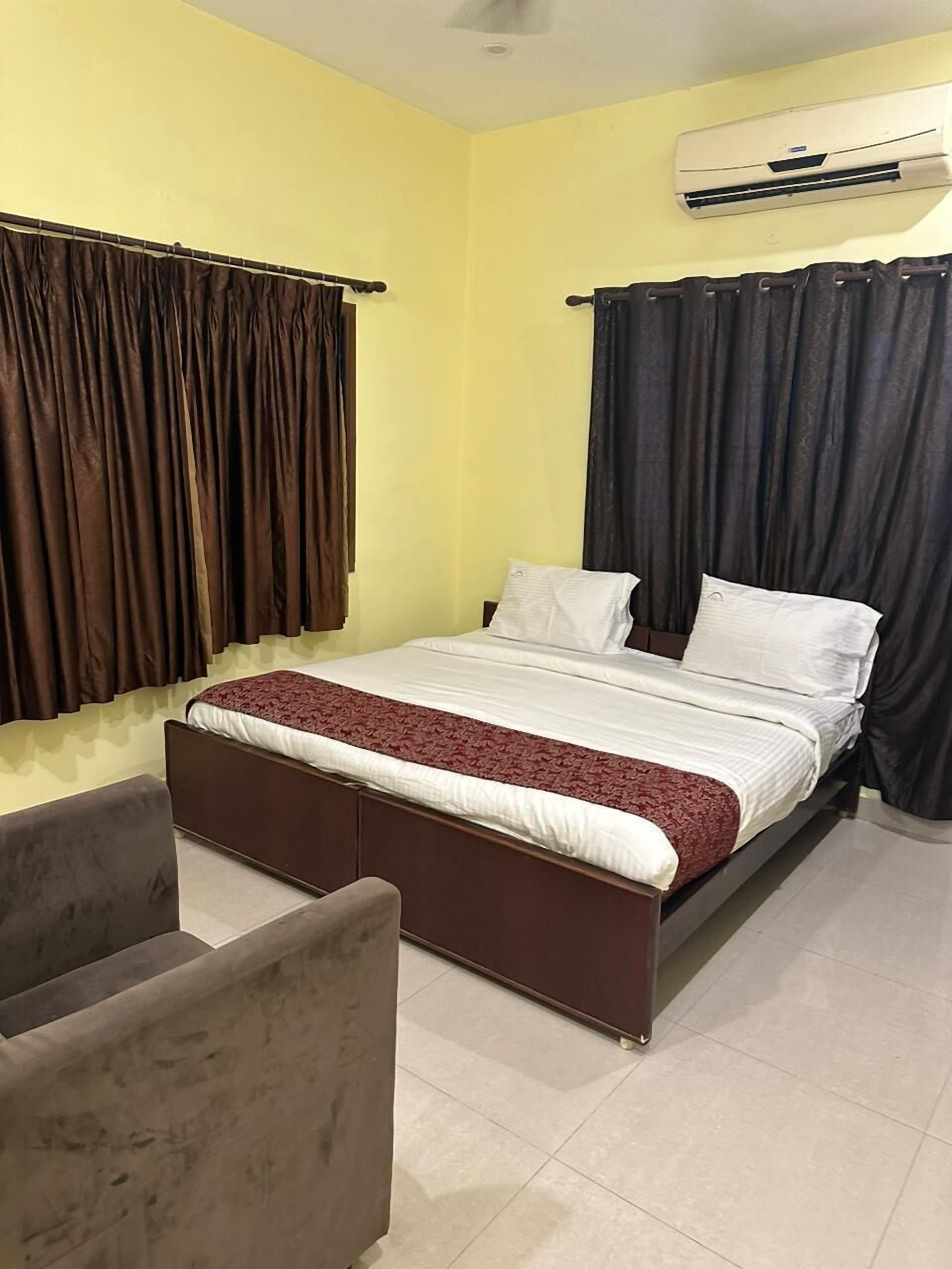 A bedroom with a bed, a chair, a window with curtains, and an air conditioning unit on the wall.