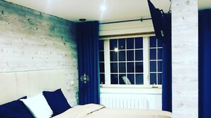A bedroom with a wooden headboard, a bed with a beige cover, blue curtains, and a window with white blinds. - Au Ptit Kakawi  (Miquelon-Langlade)
