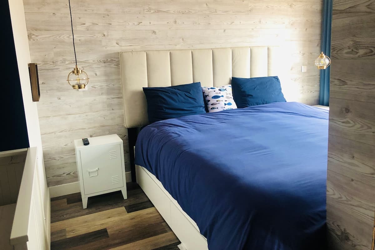 Ein Schlafzimmer mit Holzboden, einem Bett mit blauer Bettdecke, einem weißen Nachttisch und einem Beistelltisch.