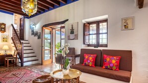 A living room with a brown sofa, colorful pillows, a round table with a vase, a staircase, and a door leading outside.