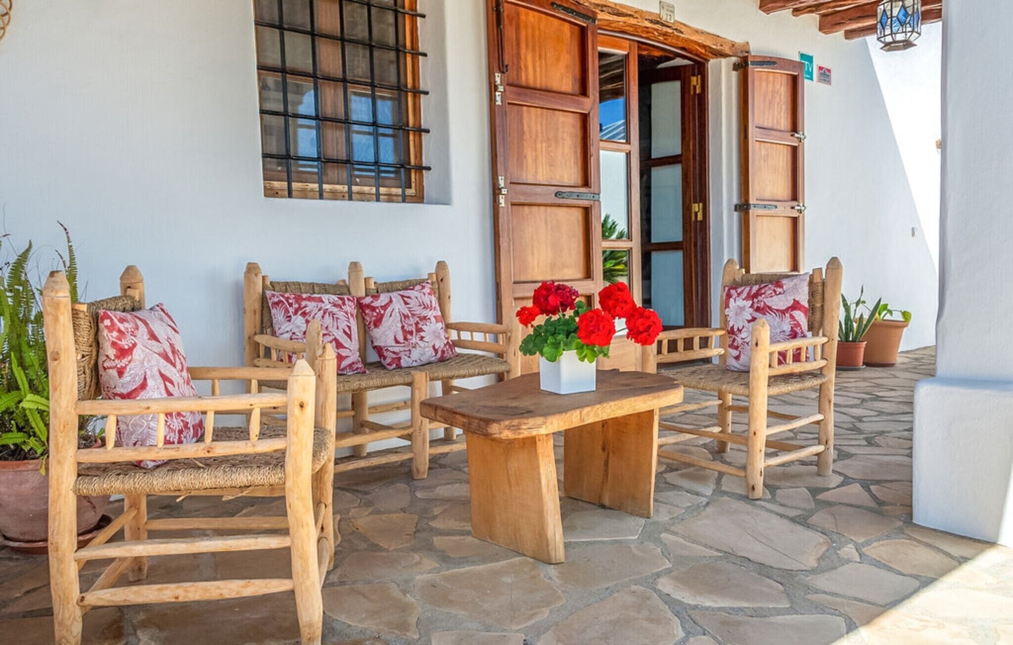 Un área de estar exterior con madera, sillas, una mesa, plantas en macetas y la entrada de un edificio.