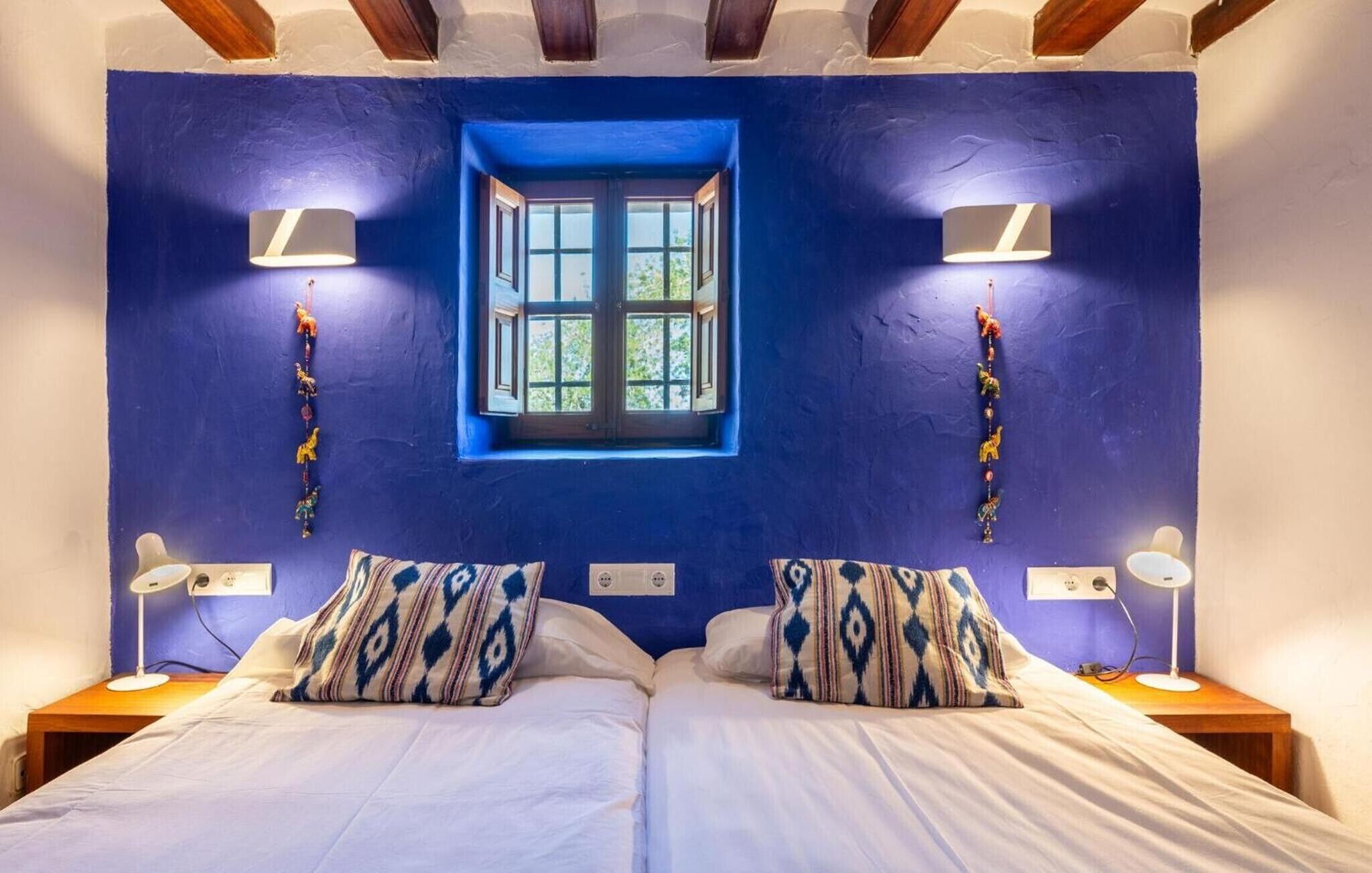 Un dormitorio con pared azul, una ventana, dos lámparas de noche y almohadones estampados.