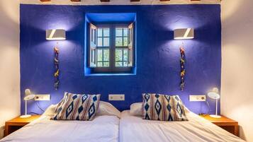 Un dormitorio con pared azul, una ventana, dos lámparas de noche y almohadones estampados.