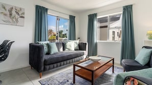 Ein modernes Wohnzimmer mit grauem Sofa, Couchtisch und Blick auf eine Wohngegend durch große Fenster mit grünen Vorhängen.