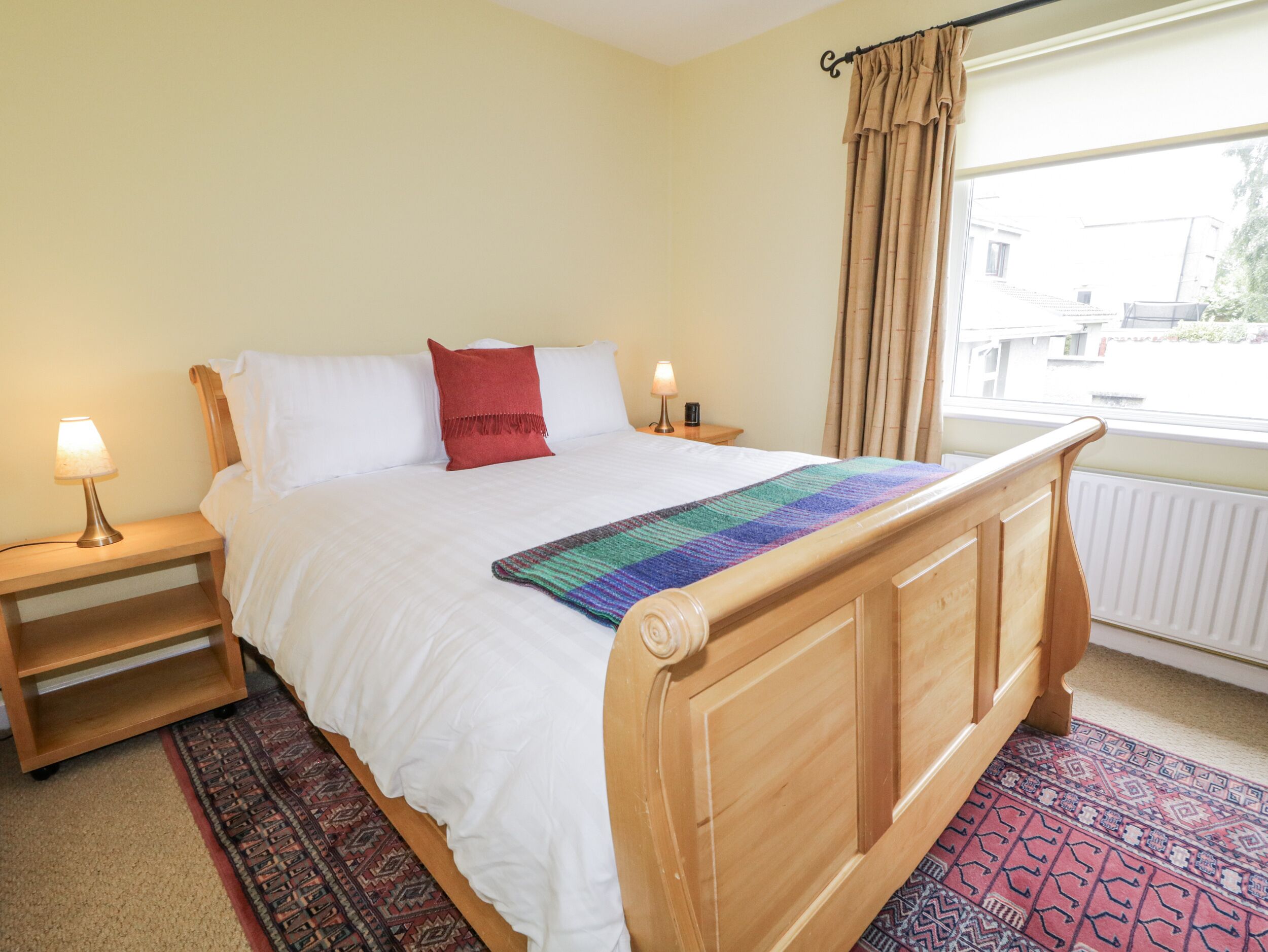 A neatly made bed with a wooden headboard, white bedding, and a colorful blanket. There are two bedside tables with lamps, a red pillow, and a window with curtains.