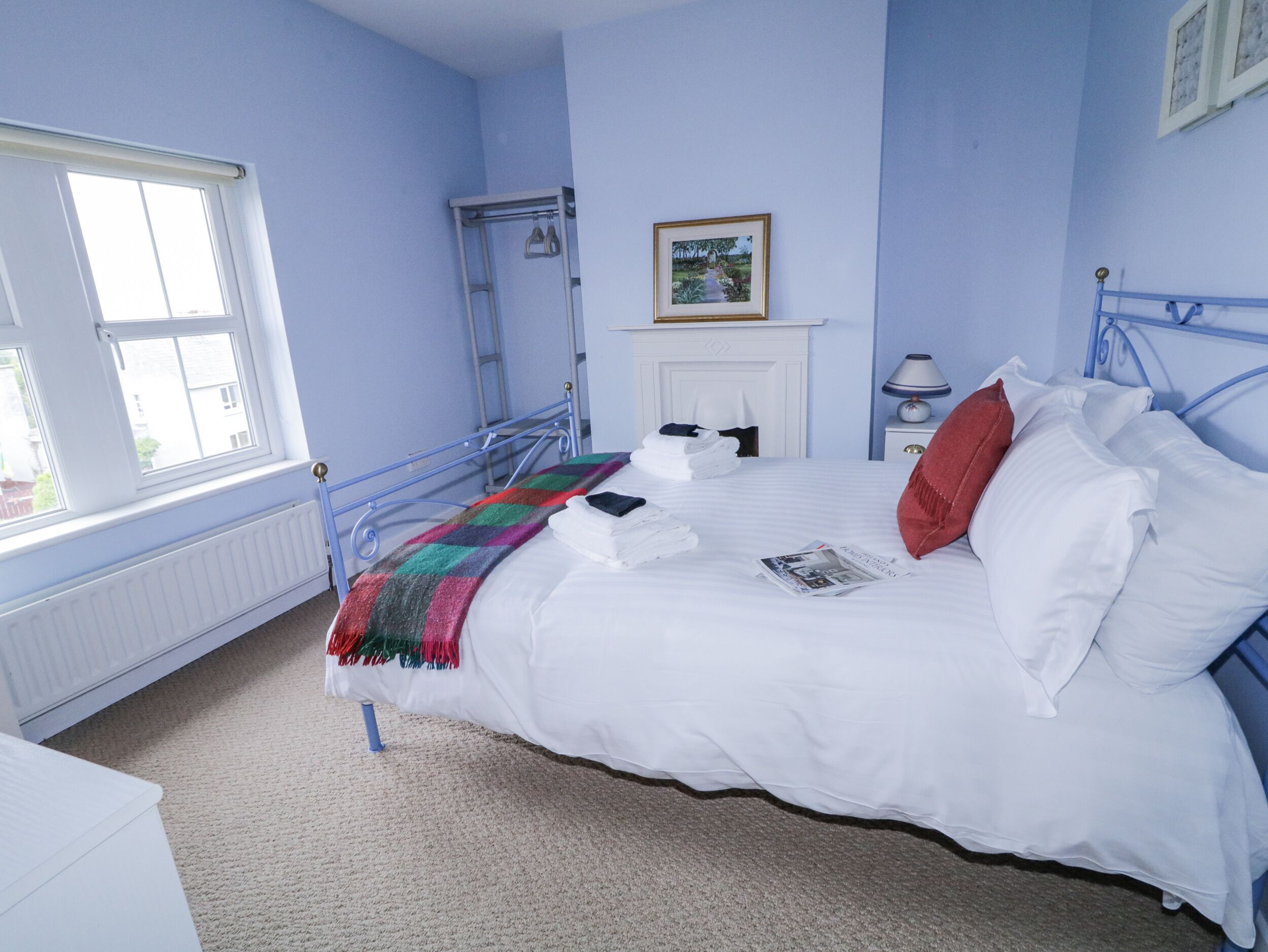 A bedroom with a bed, a window, a radiator, a framed picture, and a bedside table with a lamp.
