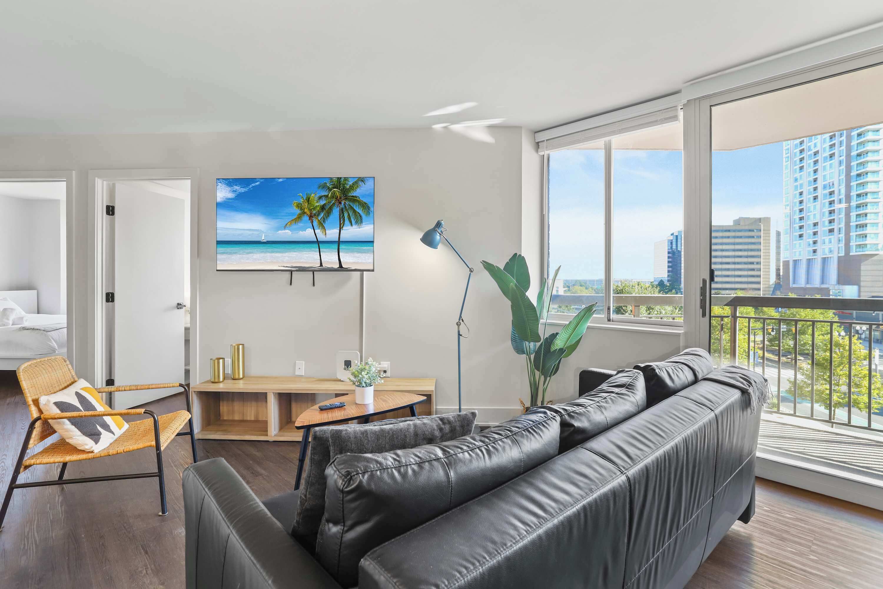 Uma sala de estar moderna com um sofá de couro preto, uma poltrona de vime, uma mesa de centro de madeira e uma grande TV exibindo uma paisagem de praia.