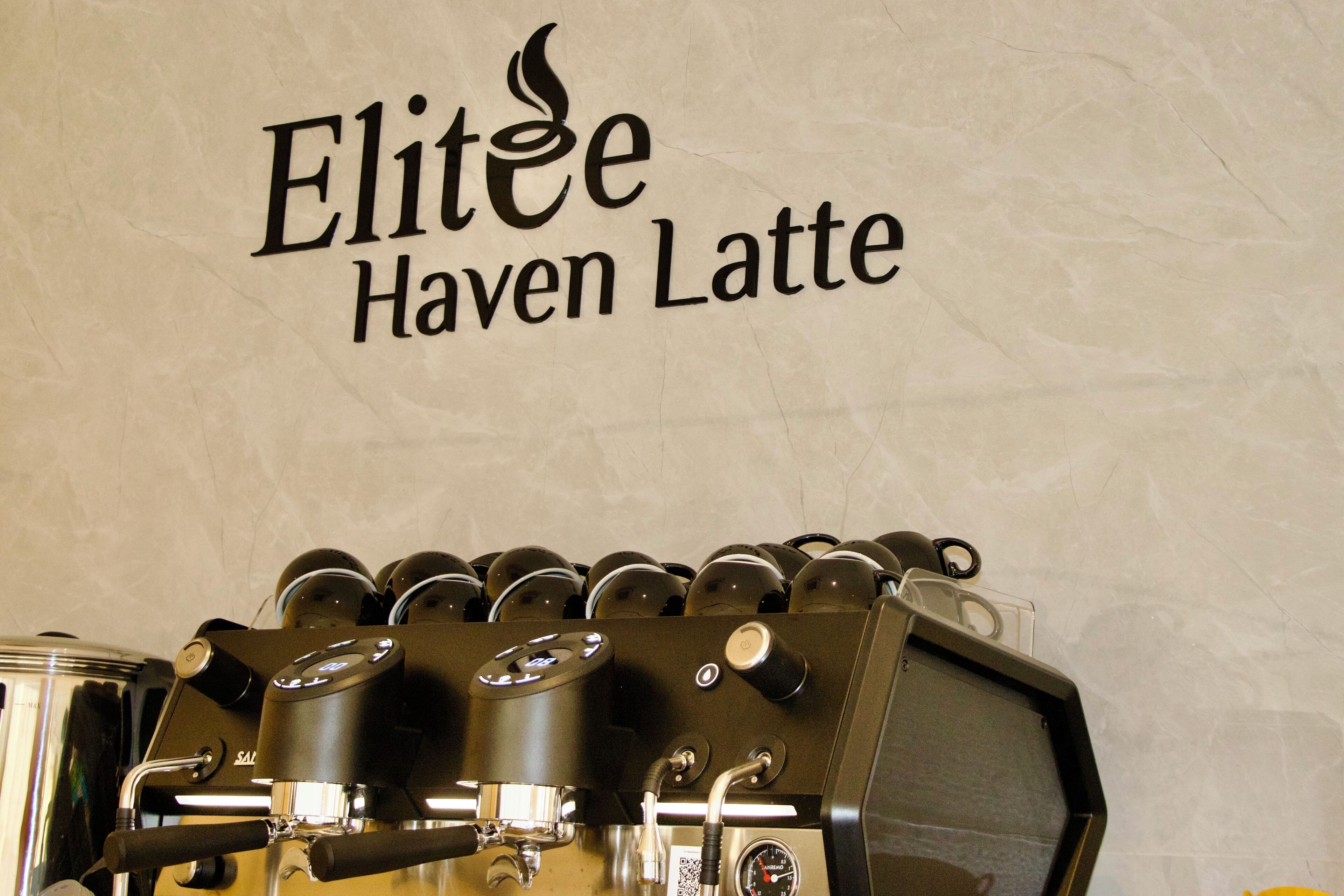 Una cafetera con varias opciones de preparación y un letrero en la pared que dice 'Elitee Haven Latte'.