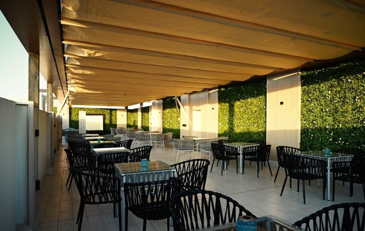 An outdoor patio area with tables and chairs, covered by a canopy, and surrounded by greenery.