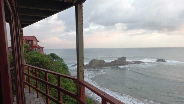 A wooden deck with a metal railing overlooking a coastal area with rocky outcrops and a building with a red roof in the background. - Apartamento con vista al mar (Tola)