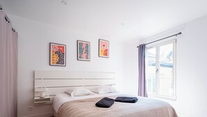 A bedroom with a white bed, a headboard, a window with curtains, and three framed floral artworks on the wall. - Le 202 Sainte Ségolène - 1 room, rénovated, heart of Metz (Metz)