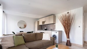 A modern living room with a sectional sofa, a round wooden coffee table, and a kitchen area with a washing machine. - Le 202 Sainte Ségolène - 1 room, rénovated, heart of Metz (Metz)