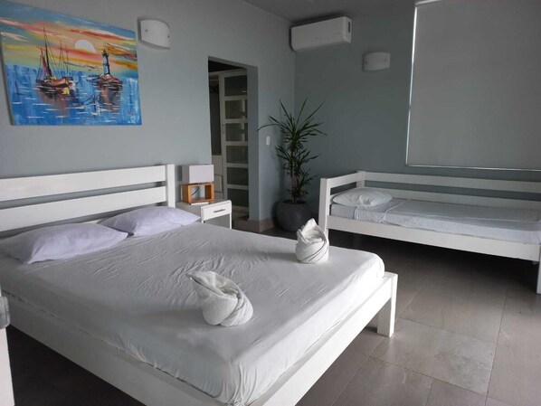 Habitación de hotel moderna con una cama grande, una cama más pequeña, una planta en maceta y un cuadro con barcos en la pared.