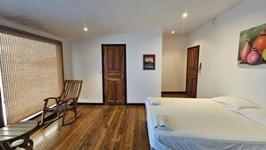 A room with a wooden floor, a bed with a white mattress and gray blanket, a rocking chair, a wooden door, and a wall-mounted picture of pottery. - Chalet con vista al Océano (Tola)