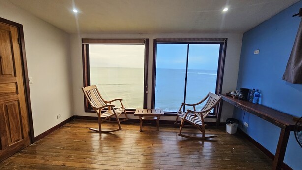 A room with wooden flooring, two rocking chairs, a small table, and a view of the ocean through large windows. - Chalet con vista al Océano (Tola)
