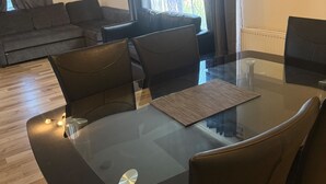 A modern living room with a glass-top dining table, black chairs, a sofa, and a view through sheer curtains. - Cozy Kirkcaldy Hideaway - 3br House (Kirkcaldy)