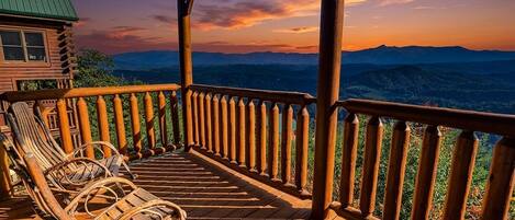 A wooden deck with a railing, two wicker chairs, and a view of mountains.