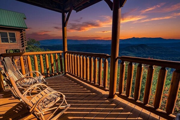 A wooden deck with a railing, two wicker chairs, and a view of mountains.
