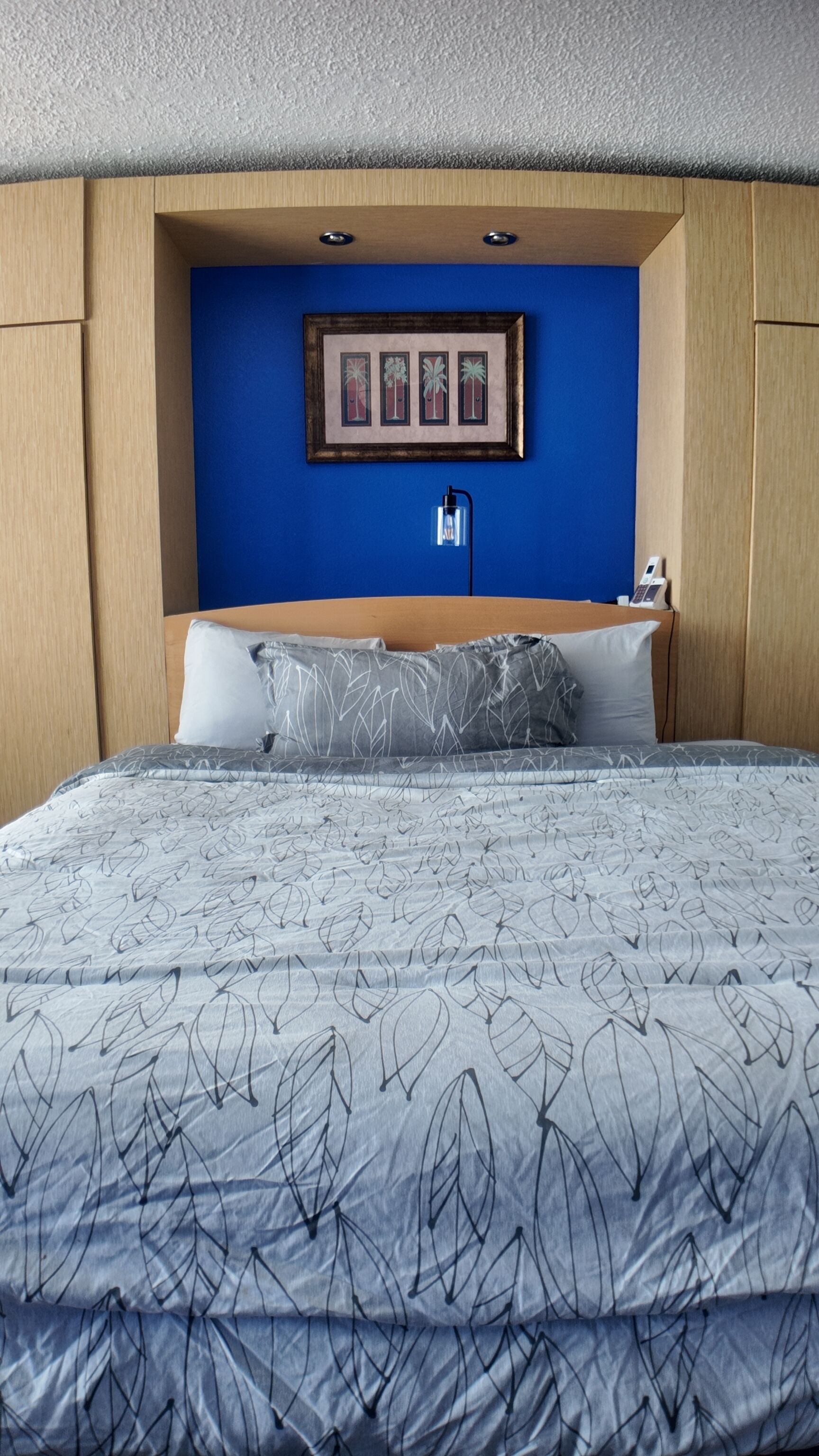 Een netjes opgemaakt bed met een dekbed met een bladpatroon, een ingelijste foto aan de muur en een nachtkastje met een lamp.