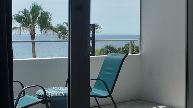 A balcony with a view of the sea, a palm tree, and a chair.