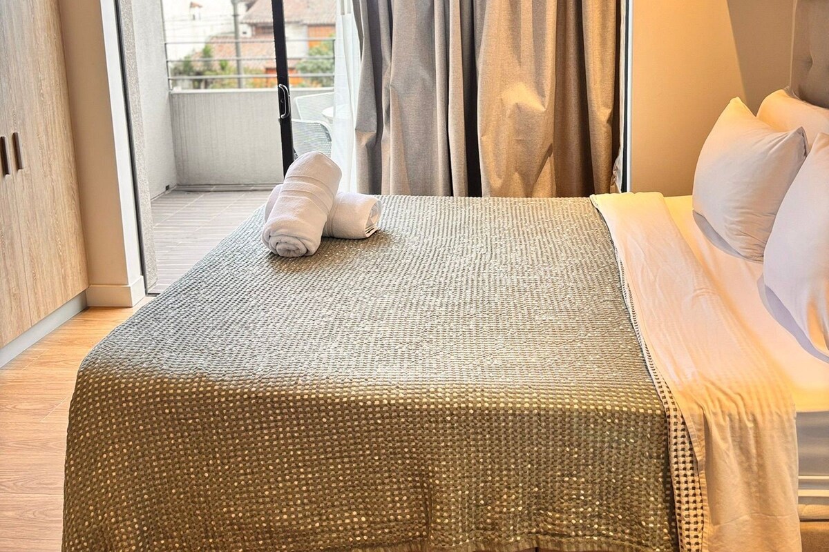A hotel room with a bed, a view through a sliding door, and a cityscape in the background.