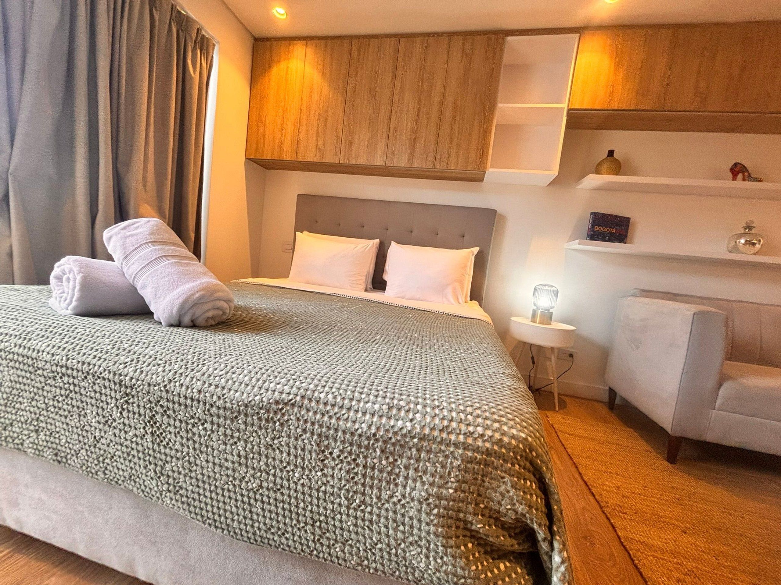 A hotel room with a bed, a nightstand, a chair, and a shelf with decorative items.