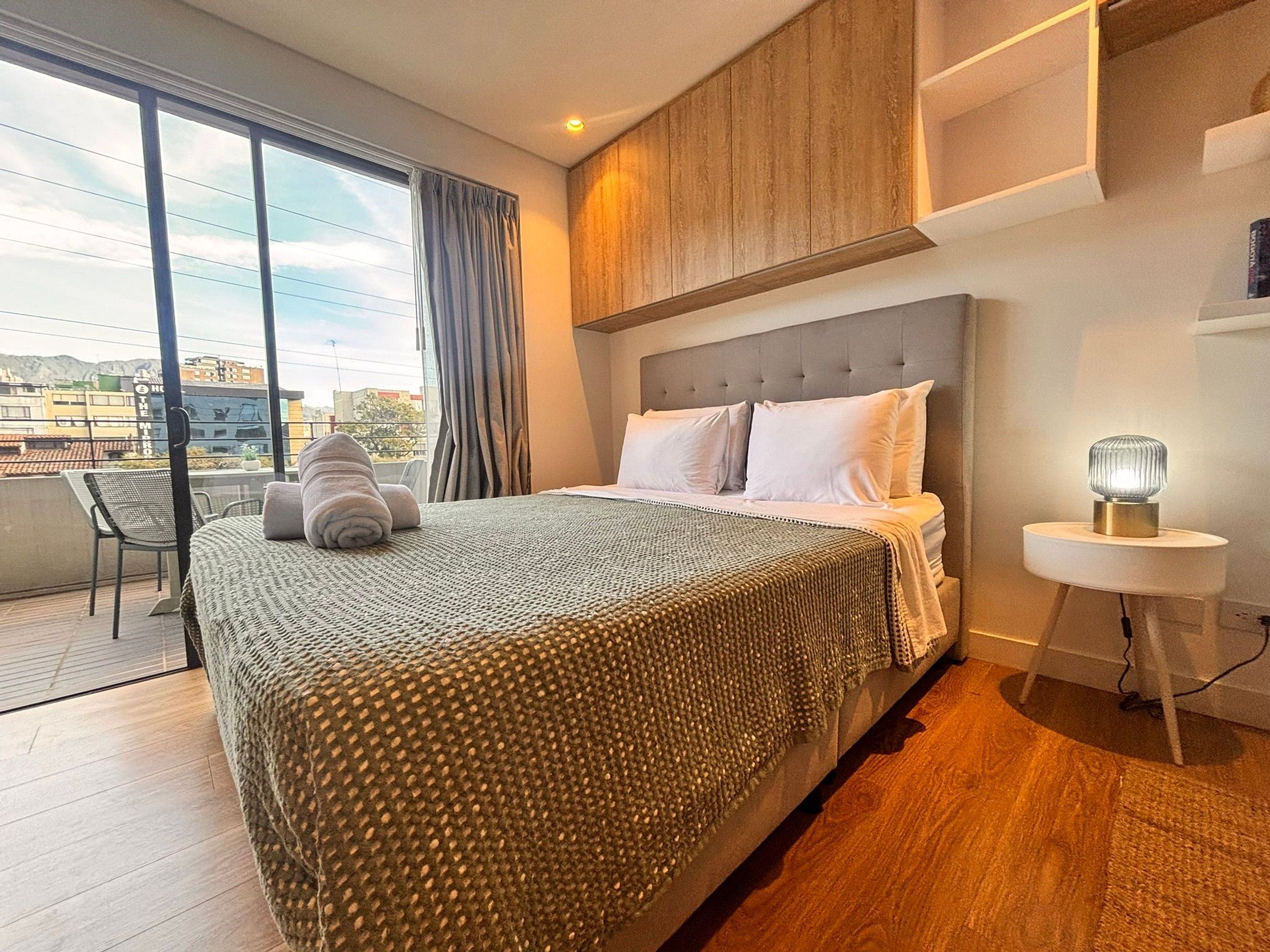 Une chambre d’hôtel dotée d’un grand lit, d’une table de chevet avec une lampe, d’une vue sur la ville depuis un balcon et d’un design moderne.