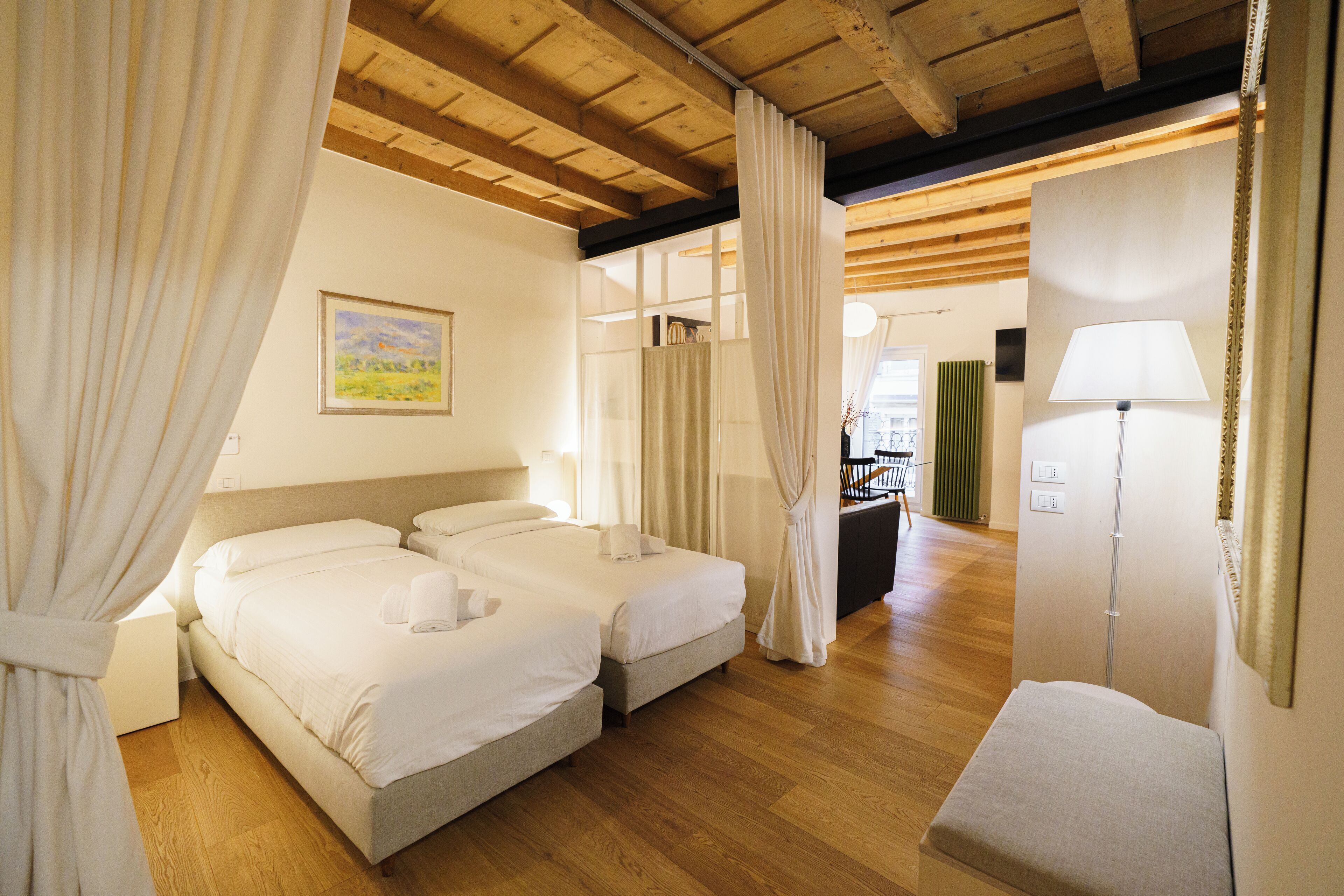 A hotel room with two beds, wooden flooring, a painting on the wall, and a lamp.