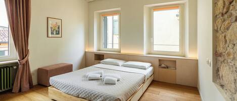 A bedroom with a wooden bed, two windows, a radiator, and a stone wall.