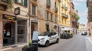 Una calle con un SUV blanco estacionado en la banquina, edificios con balcones y un cartel que dice 'DOPPIO MALTO'.