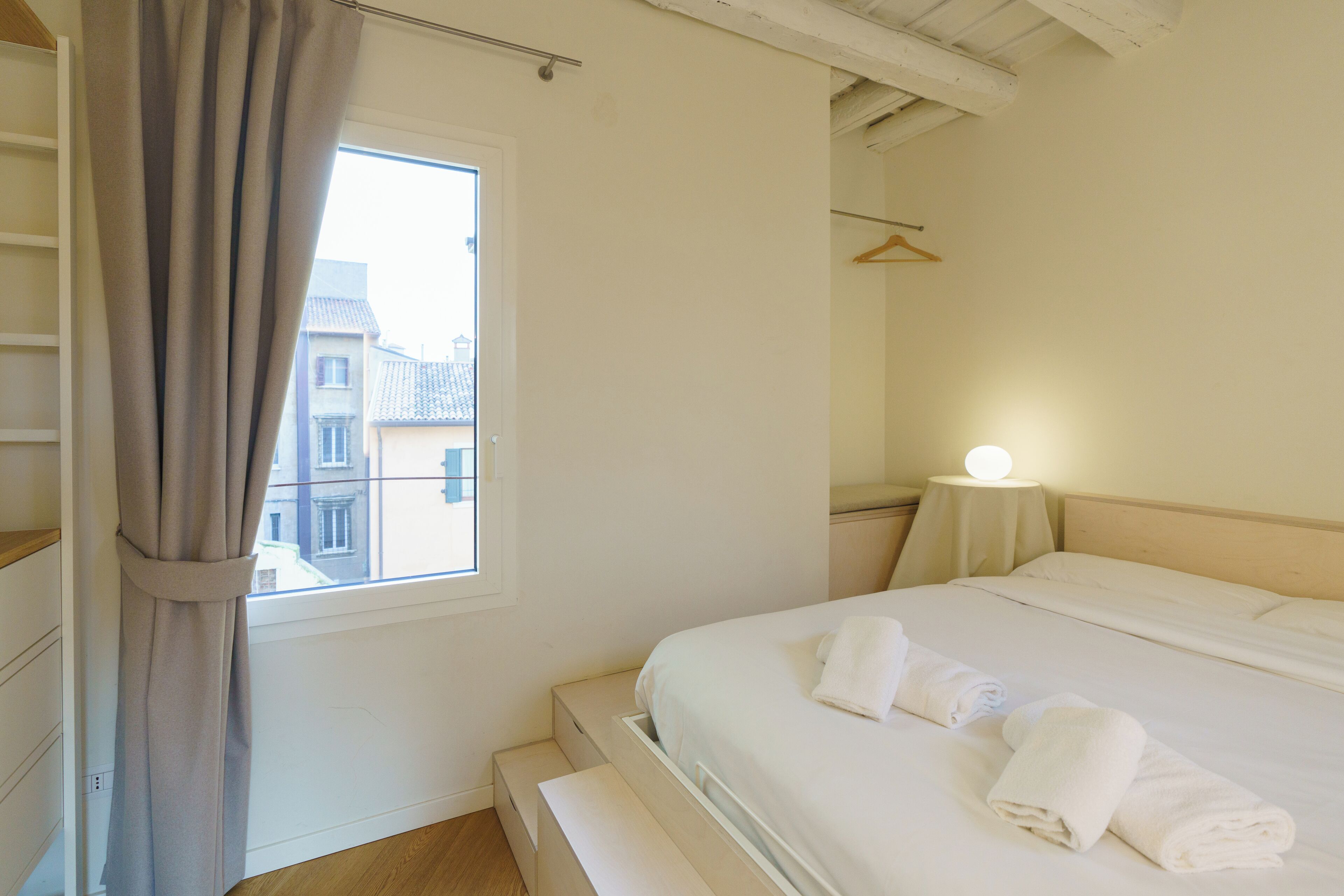 Een slaapkamer met een bed, een nachtkastje met een lamp, een raam met gordijnen en uitzicht op gebouwen.