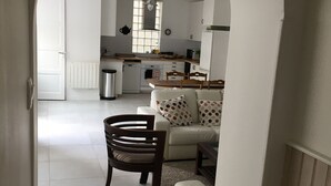 Un vaste espace de vie avec un canapé blanc et une chaise en bois, une cuisine en arrière-plan et une porte blanche donnant sur une autre pièce.