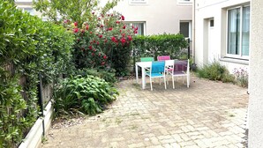 Une cour avec une terrasse dallée, une petite table entourée de chaises, ainsi qu’une variété de plantes et de fleurs.