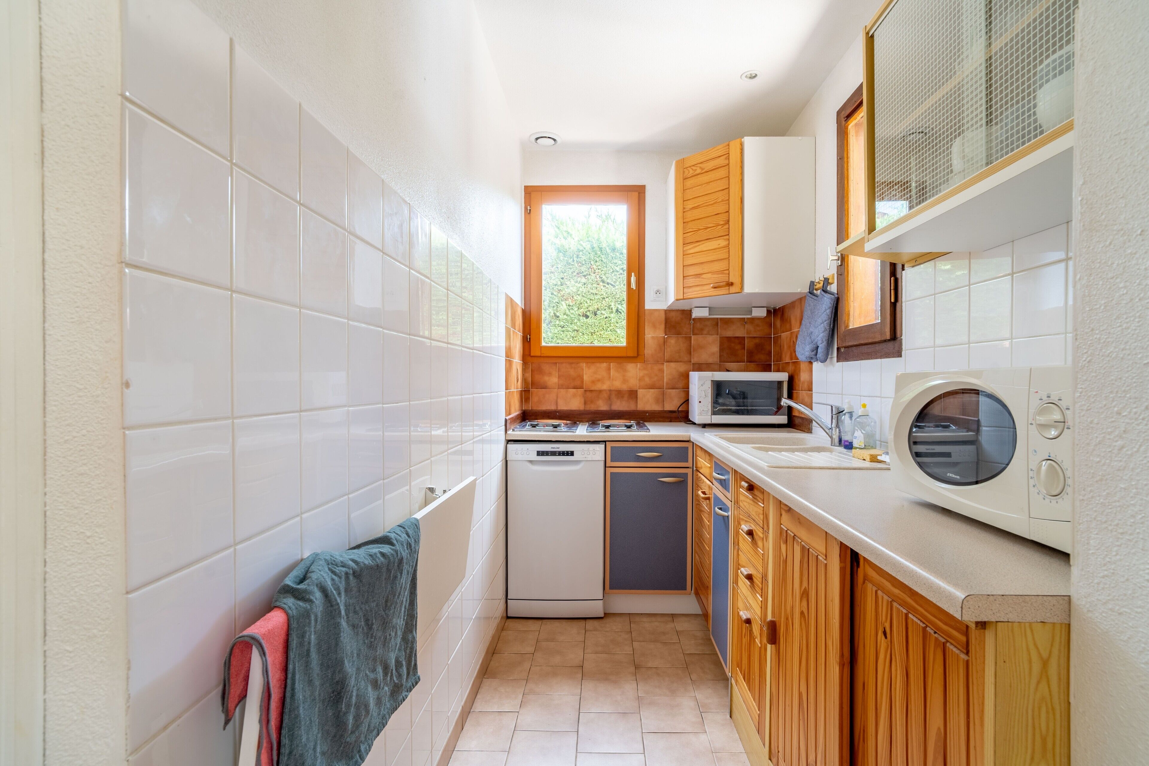 A compact kitchen with white tiles, a dishwasher, a microwave, wooden cabinets, and a window.