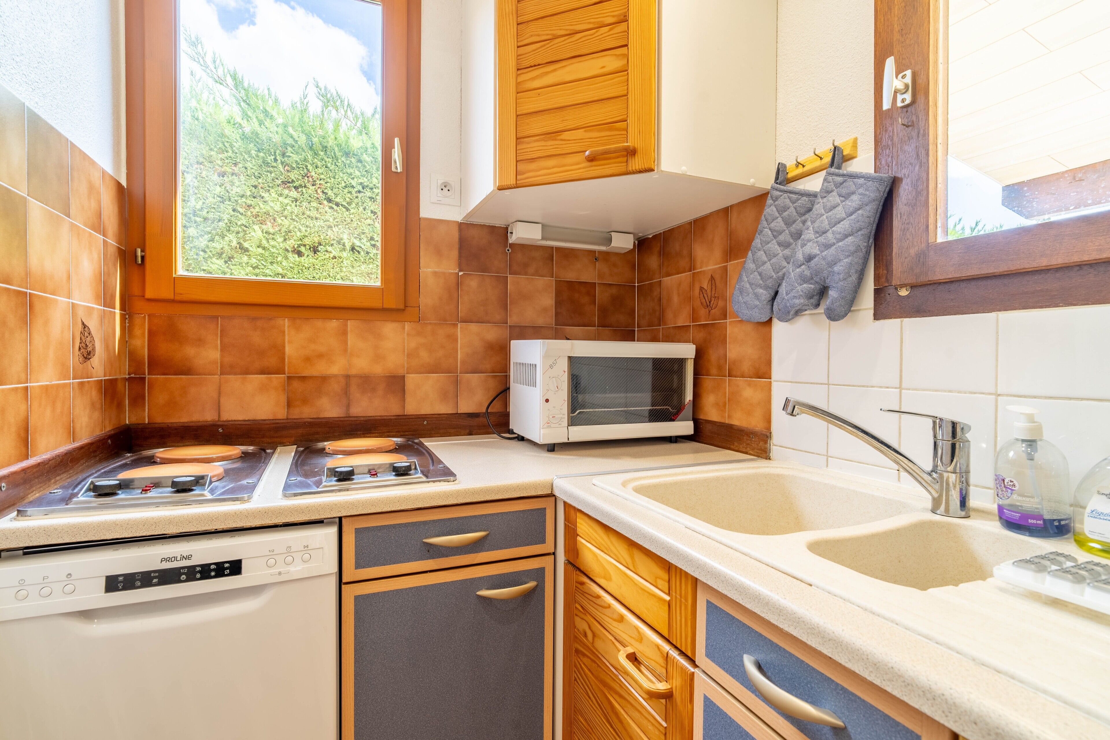 A kitchen with a stove, microwave, sink, wooden cabinets, and a window with a view of greenery.