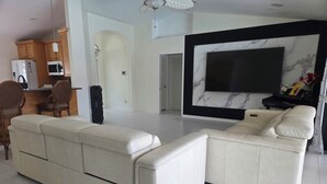 A modern living room with a large sectional sofa, a flat-screen TV mounted on a marble wall, and a kitchen area with wooden cabinets and appliances.