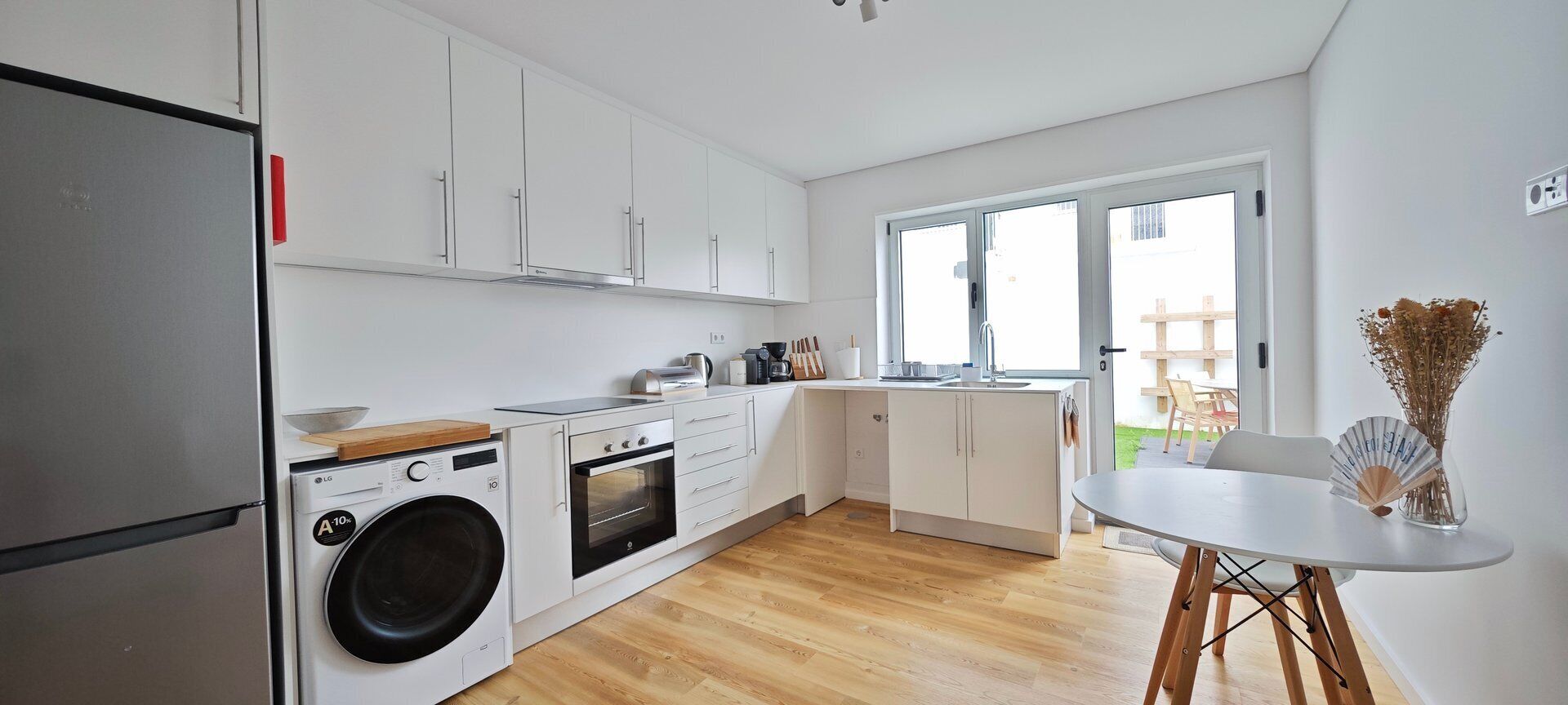 Een moderne keuken met witte kasten, een wasmachine, een oven en een eethoek met een tafel en stoelen.