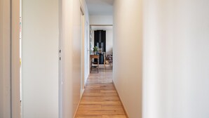 Un couloir étroit doté d’un plancher en bois, de murs blancs et d’une porte au fond, qui donne sur une pièce contenant une table et des chaises.