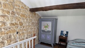 A bedroom with a stone wall, wooden beams, a bed with striped bedding, a nightstand with a lamp, and a dresser with a floral decoration.