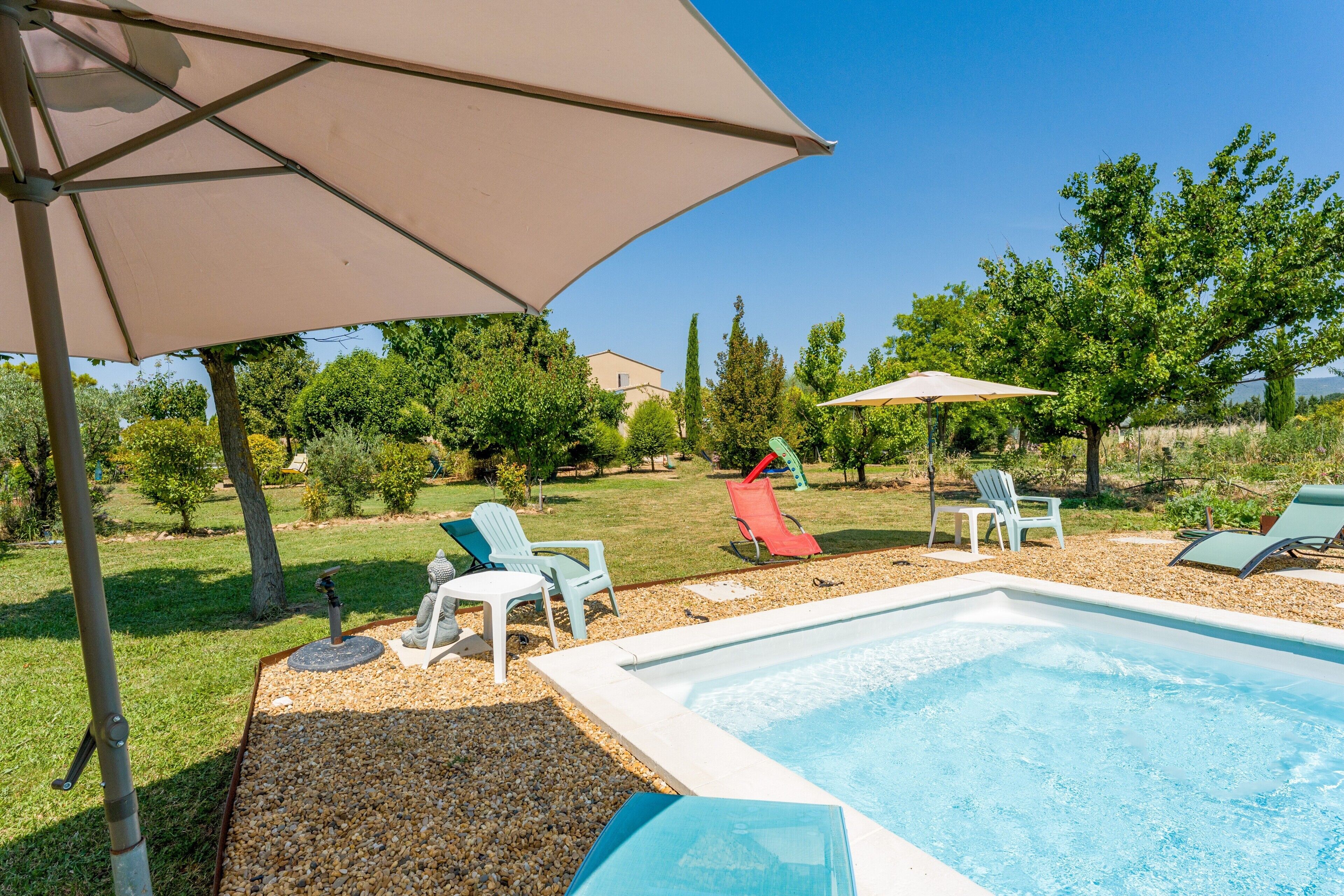 Une piscine entourée de chaises longues et de parasols, elle-même bordée d’un jardin avec des arbres et une pelouse.