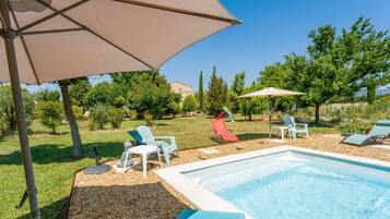 Une piscine entourée de chaises longues et de parasols, elle-même bordée d’un jardin avec des arbres et une pelouse.