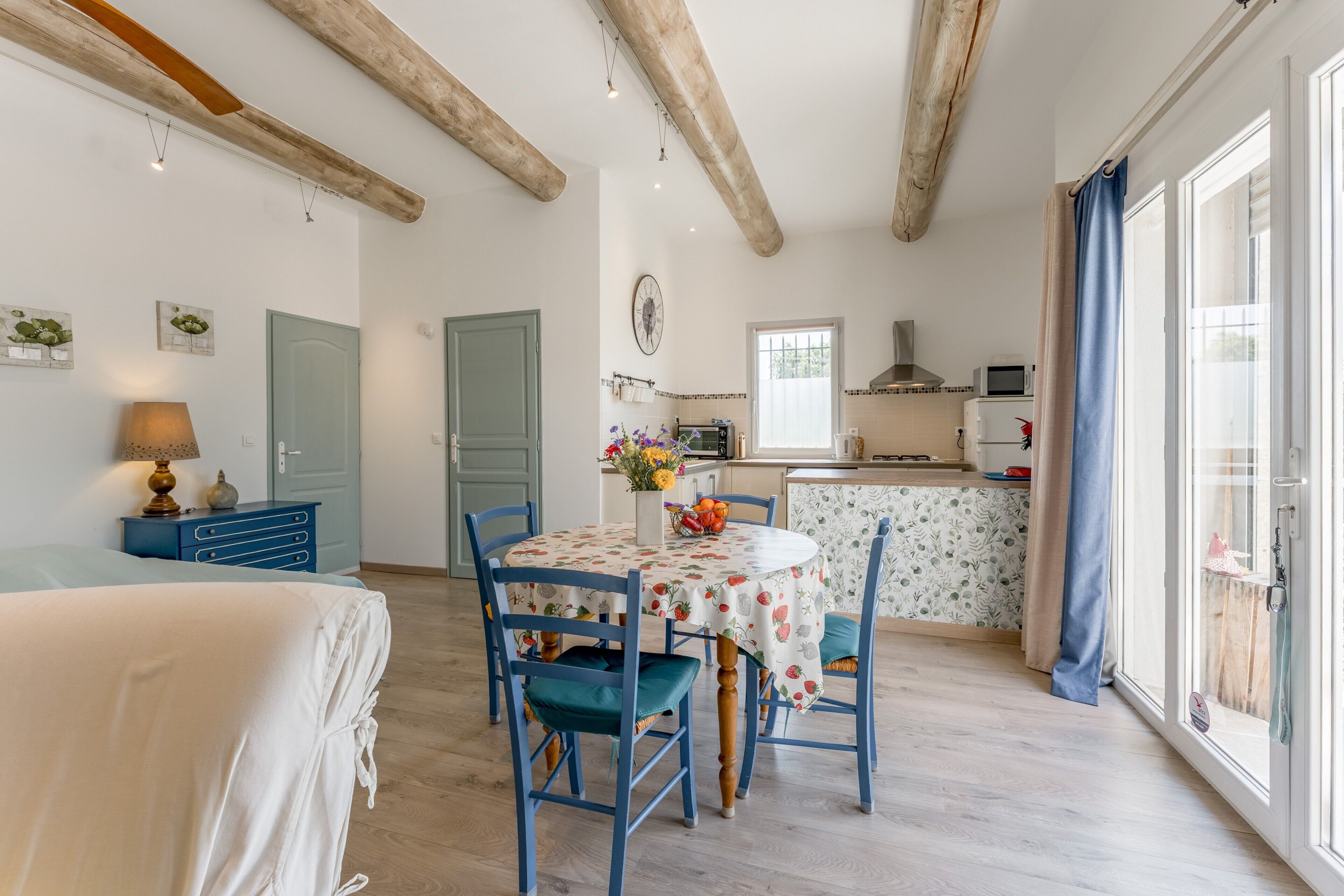 A room with a bed, a dining table with chairs, a kitchen area, and a large window with curtains.