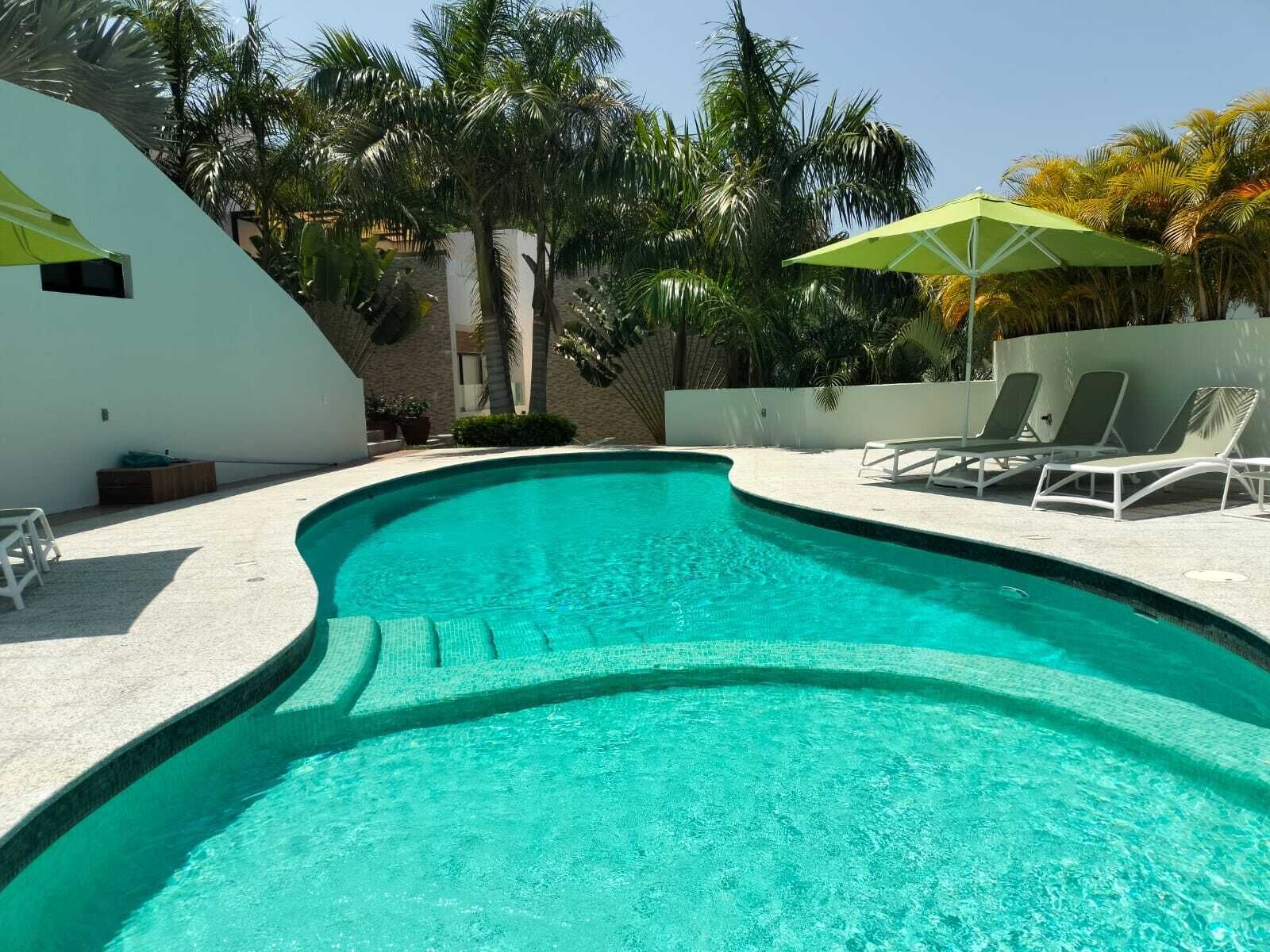 Une piscine à bord incurvé, entourée de chaises longues blanches et d’un parasol vert.